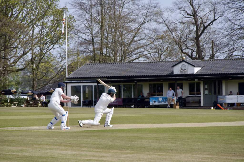 Cricketer playing a front-foot shot during a match with wicketkeeper and pavilion in the background